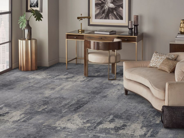 Modern living room corner with a beige sofa, patterned throw pillow, gold-and-brown side table, framed wall art, tall vases, and a blue-gray textured carpet covering the floor.